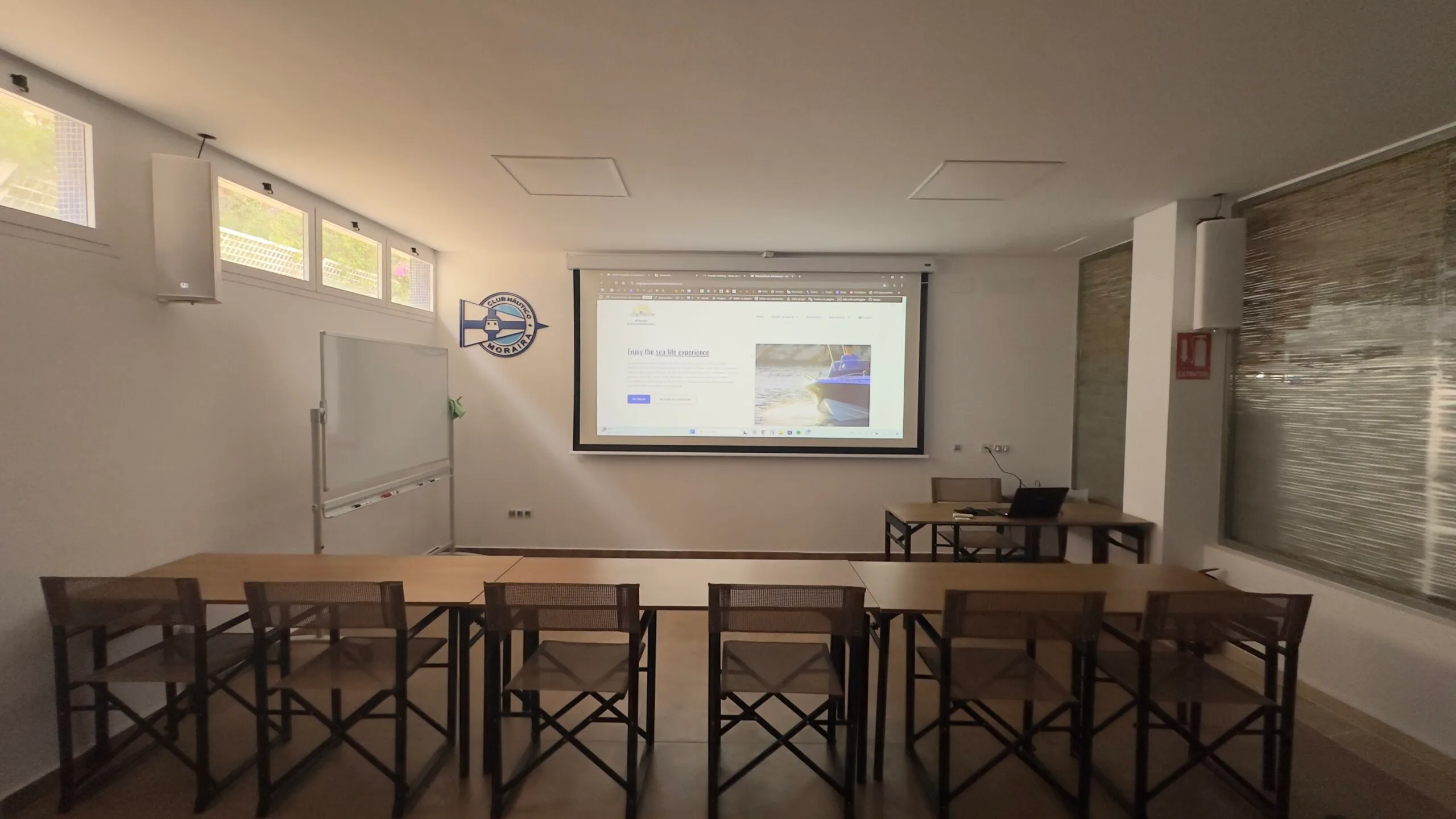 Aula con mesas y sillas plegables frente a una pantalla que proyecta una página web, con una pizarra blanca a un lado