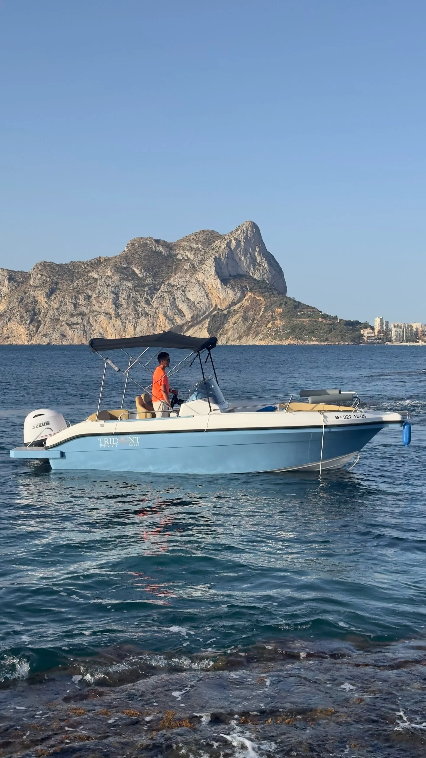 Embarcación fondeada junto a las rocas, con el Peñón de Ifach al fondo y una persona al timón