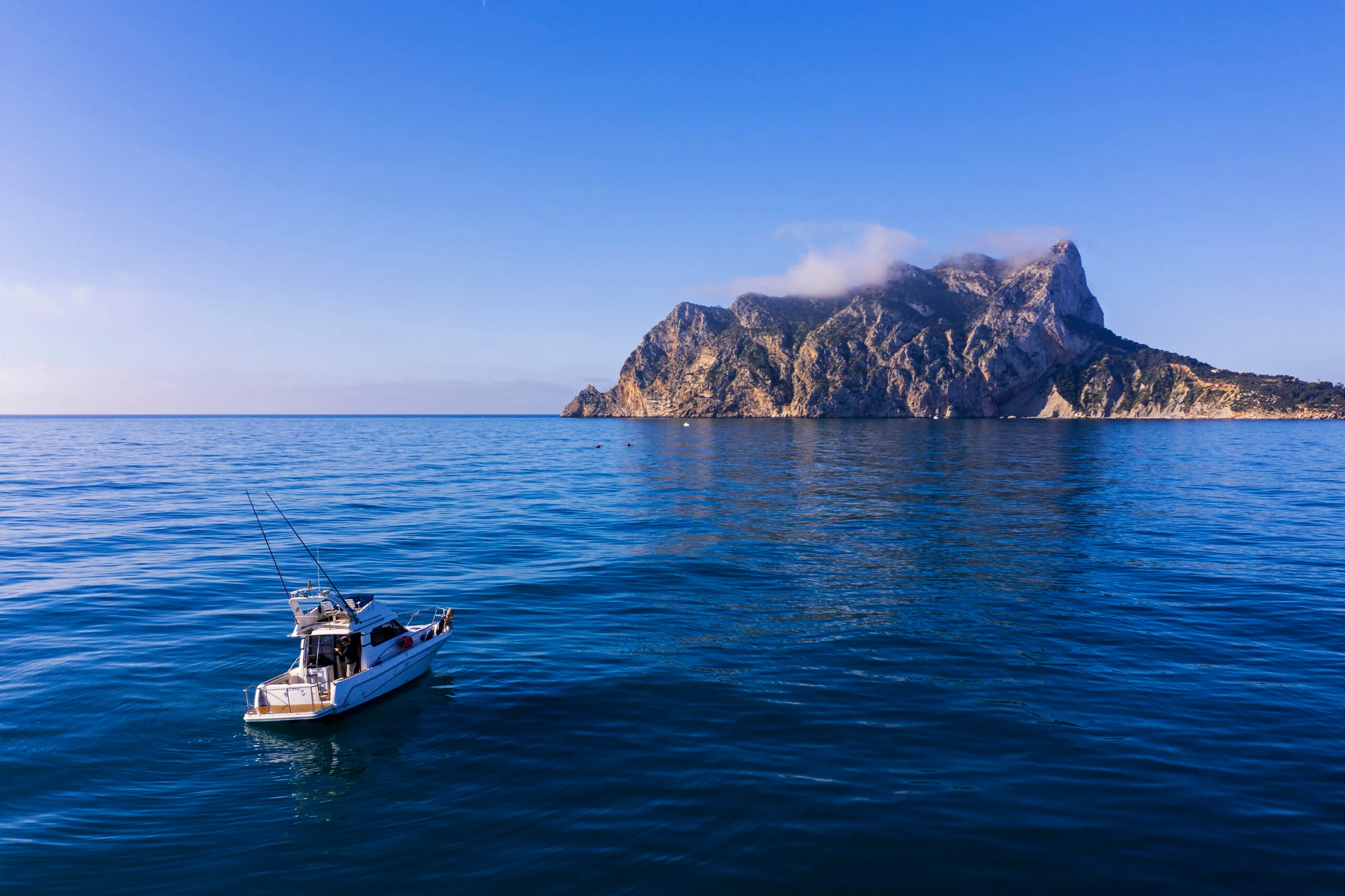 Embarcación con cañas de pesca navegando en un mar en calma, con el Peñón de Ifach al fondo