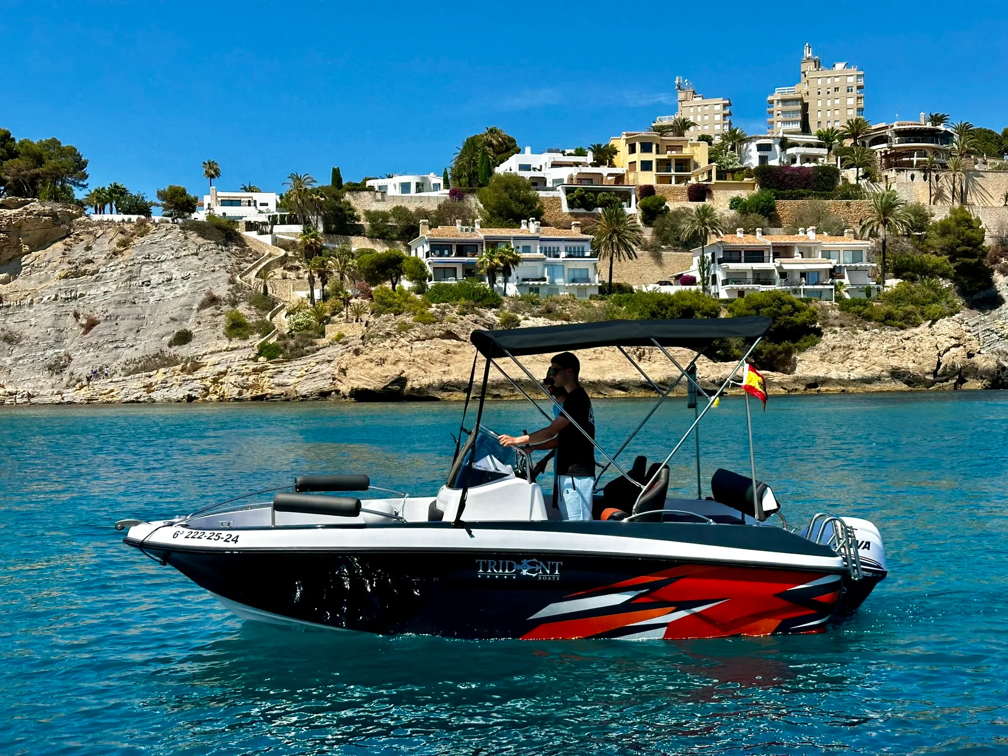 Embarcación pequeña a motor, negra y naranja, con el patrón al timón frente a la costa